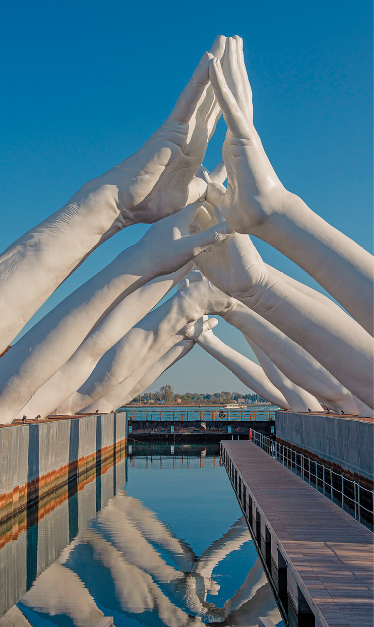 Escultura. Em um espaço aberto, próximo a um rio, com destaque para uma escultura de diversos antebraços estendidos para o alto e, um de cada lado, eles entrelaçam as mãos ao alto, formando um corredor sobre eles.