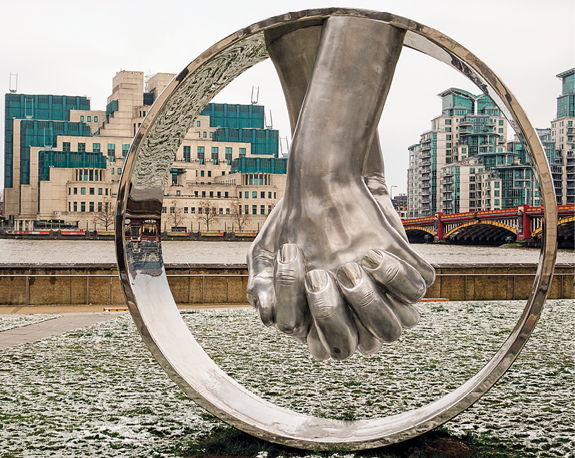 Escultura. Escultura de duas mãos dadas para baixo dentro de uma estrutura anelar prateada. Ao fundo, vista de um rio, uma ponte e alguns prédios.