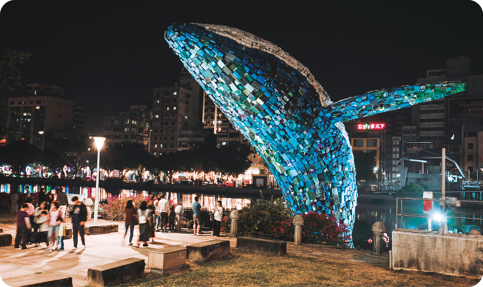 Escultura. Vista noturna de algumas pessoas em uma praça próximas a uma escultura gigante de um robusto animal marinho com corpo azul brilhante e nadadeiras longas.