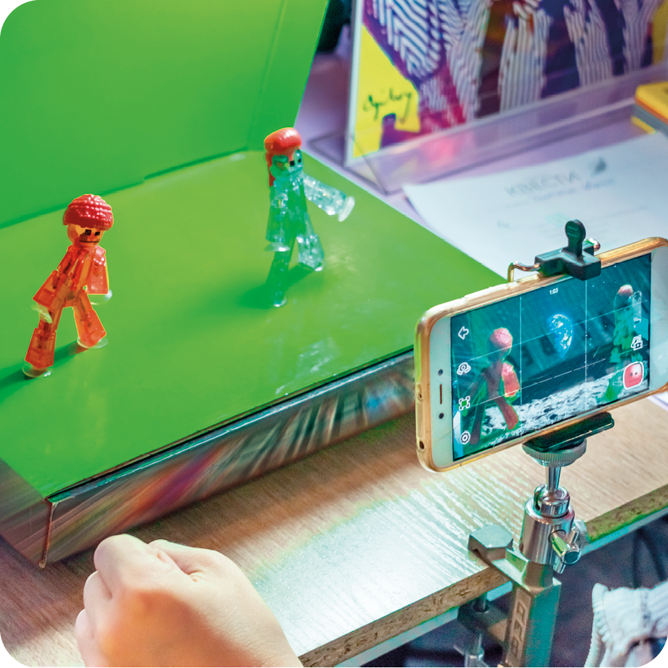 Fotografia. Destaque para um celular sobre um suporte gravando a cena de dois bonecos dispostos sobre uma plataforma com fundo verde.