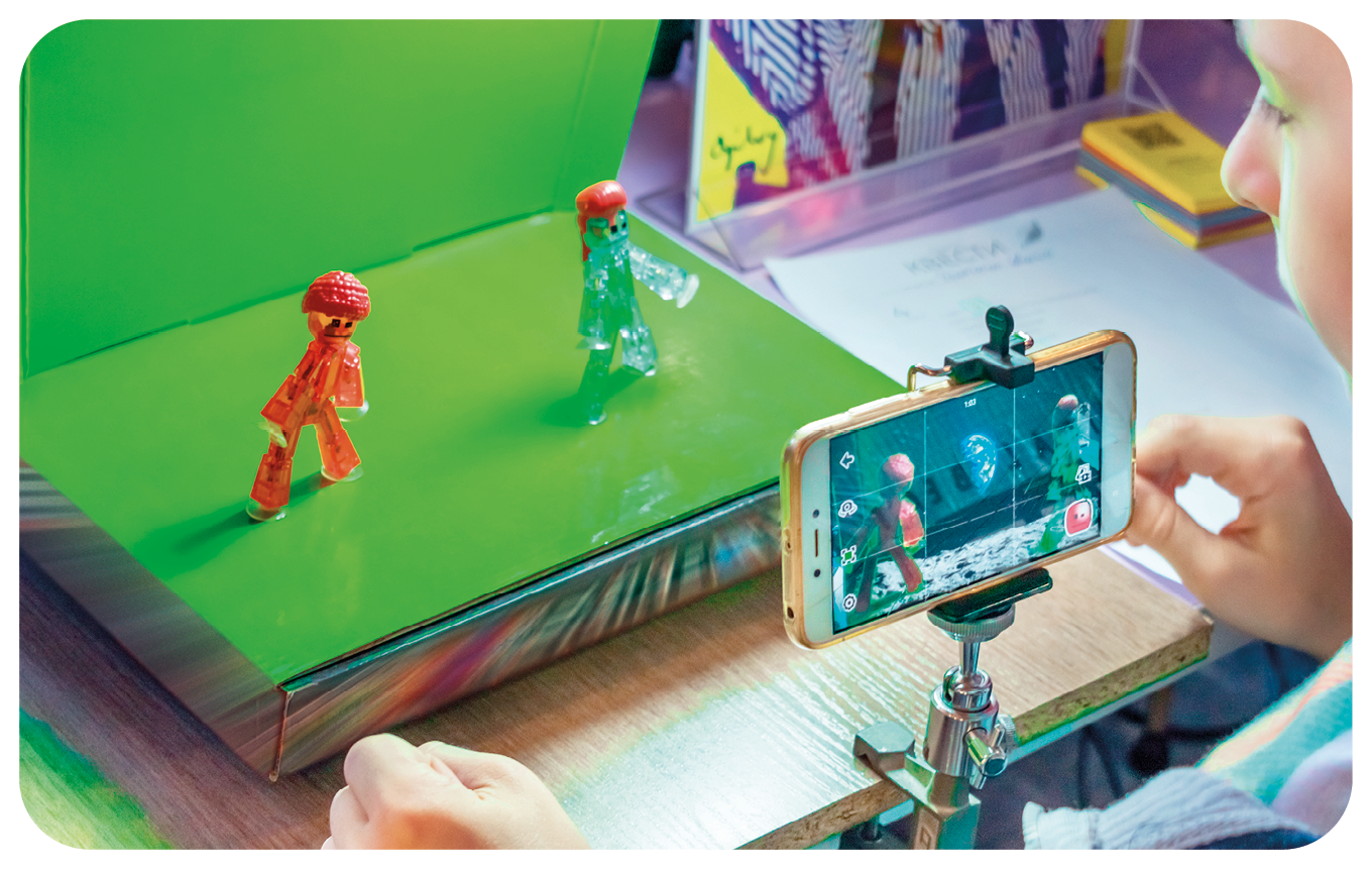 Fotografia. Destaque para um celular sobre um suporte gravando a cena de dois bonecos dispostos sobre uma plataforma com fundo verde.