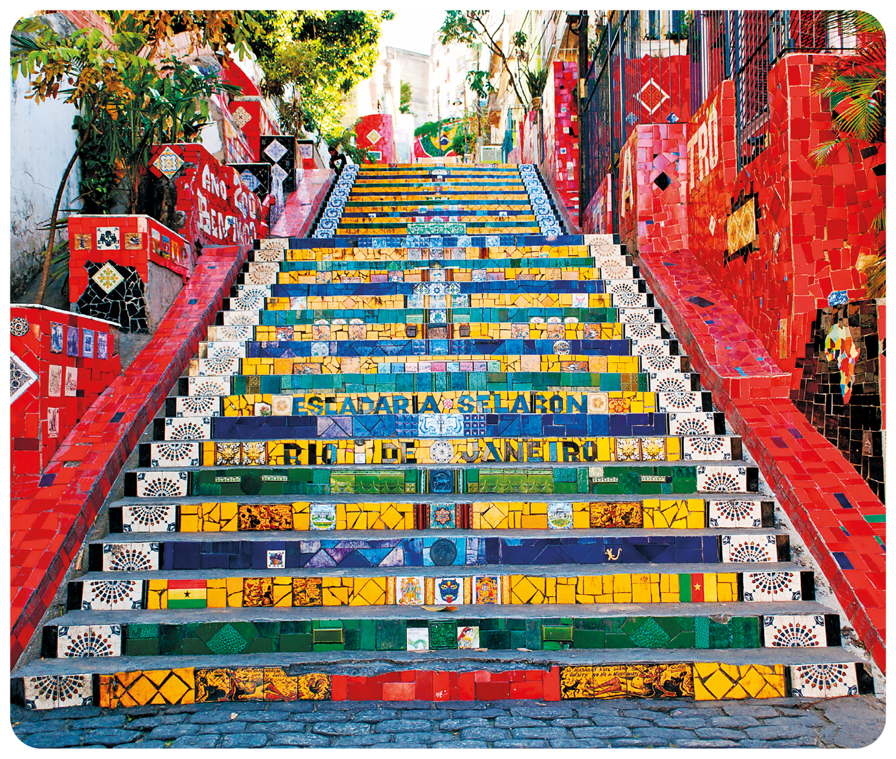 Fotografia. Plano aberto de uma escadaria com degraus compostos de ladrilhos coloridos, em tons de amarelo, azul, verde e branco e laterais com ladrilhos avermelhados formando uma composição de mosaico. Nos degraus centrais, está escrito: ESCADARIA SELARON. RIO DE JANEIRO.