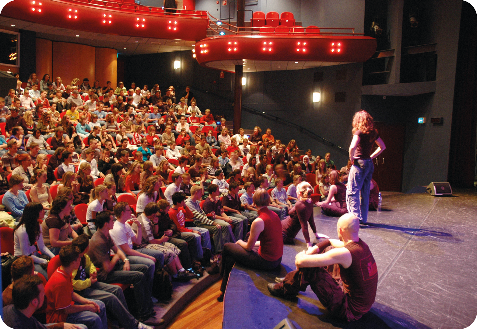 Fotografia. Vista lateral do interior de um teatro lotado. A plateia observa um grupo de seis pessoas paradas em um pequeno palco; cinco estão sentadas e uma em pé com as mãos na cintura. Na parte superior, vista parcial de um camarote com cadeiras vermelhas vazias.