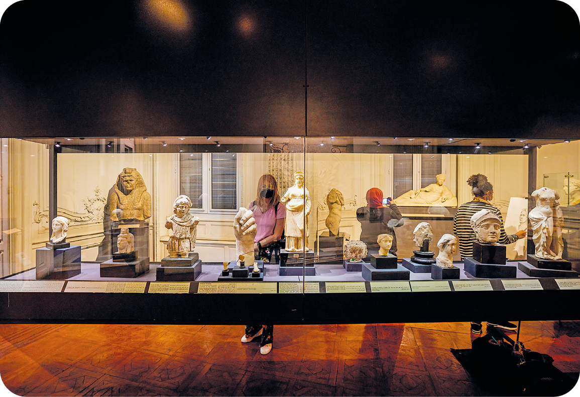 Fotografia. Em uma sala com pouca iluminação, há uma galeria com parede de vidro que apresenta esculturas retratando alguns bustos e outros objetos. Entre as paredes de vidro da galeria, alguns visitantes caminham e observam as obras.