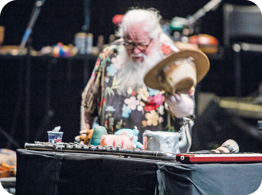 Fotografia. Vista frontal e da cintura para cima de um homem grisalho, de camisa florida, óculos de grau e barba volumosa. Ele está segurando um chapéu e diante de uma mesa com diversos objetos e instrumentos.