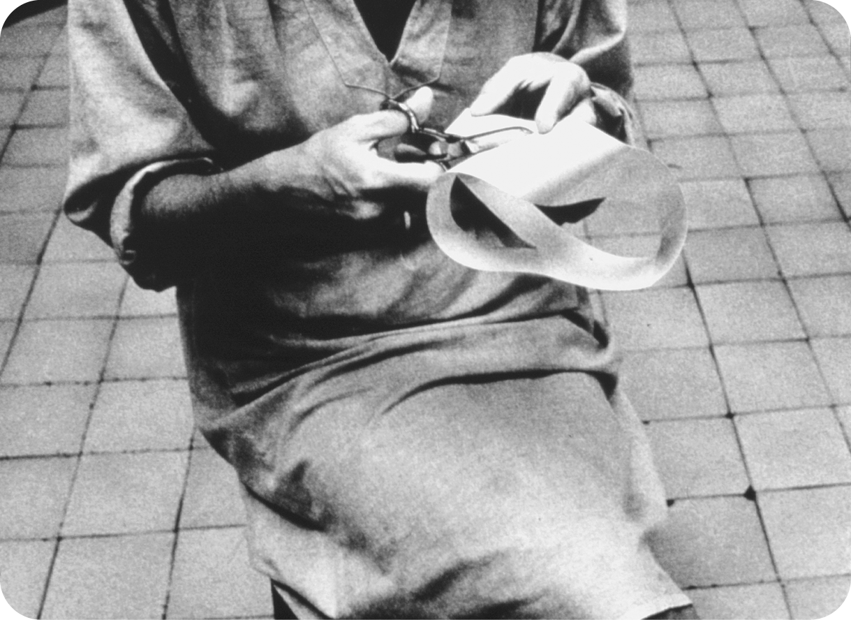 Fotografia em preto e branco. Uma mulher vista do tronco até os joelhos está sentada  com as pernas cruzadas e usando vestido largo. Ela recorta uma fita grossa branca.