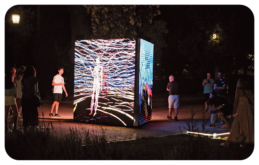 Fotografia. Durante a noite, em ambiente aberto, um grupo de pessoas está ao redor de um grande cubo composto por painéis com imagens luminosas.
