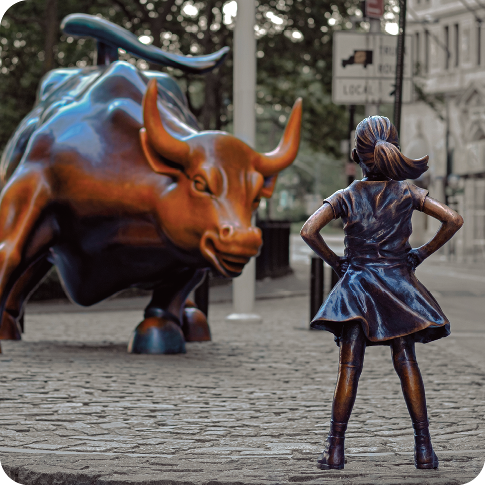Fotografia. Destaque de uma via que apresenta duas estátuas de bronze: uma menina e um touro. De um lado, vista de costa, está uma menina de vestido parada com as mãos na cintura diante de um touro visto de frente com as patas flexionadas. Ao fundo, estão alguns prédios.