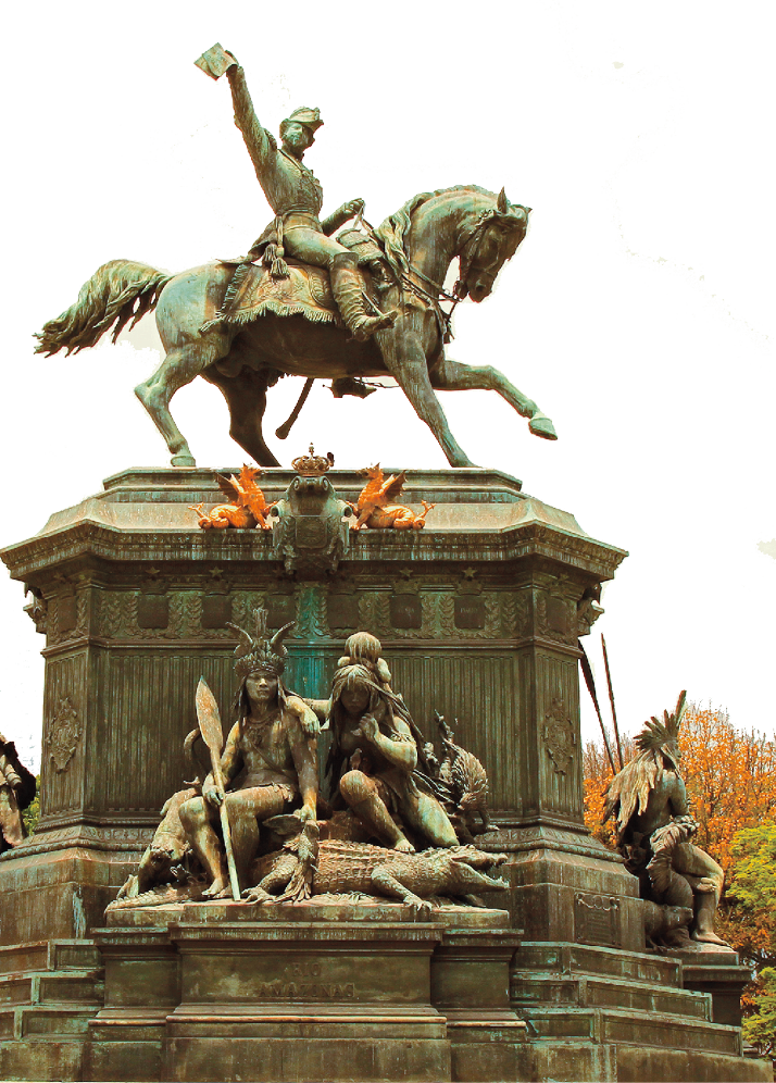 Fotografia. Destaque de uma estátua robusta que apresenta pessoas indígenas sentadas na base ao lado de animais e plantas e um homem de farda a cavalo no topo. Ele está com um dos braços levantados e olha para o lado; o cavalo está com a pata esquerda erguida.