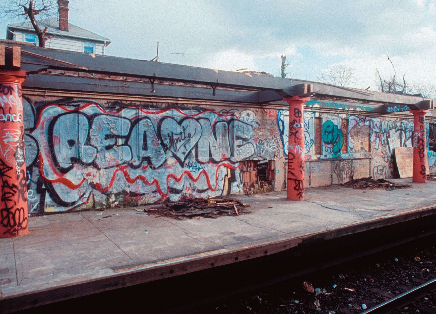 Fotografia. Destaque de uma plataforma de estação de trem que apresenta colunas e parede com grafite.
