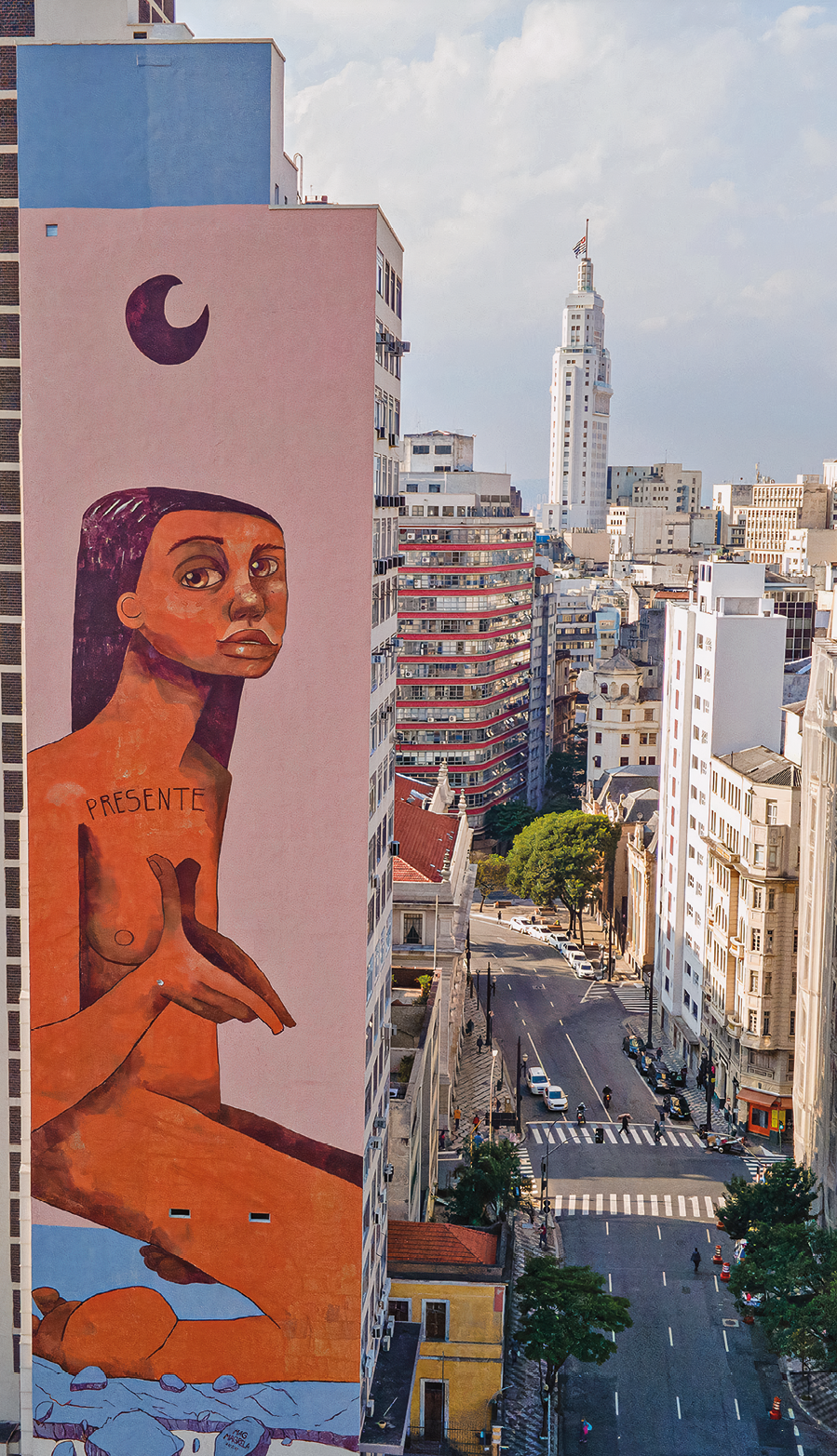 Fotografia. À esquerda, destaque de um edifício com grafite na parede lateral que apresenta o desenho de uma mulher nua sentada sobre as pernas e uma meia-lua na parte superior. À direita, está uma ampla via e prédios.