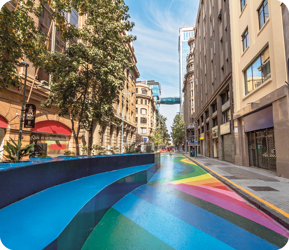 Fotografia. Destaque de um piso em uma via pública com faixas retas e curvas coloridas, em tons de azul, verde, rosa, vermelho e amarelo. Ao redor, há prédios.