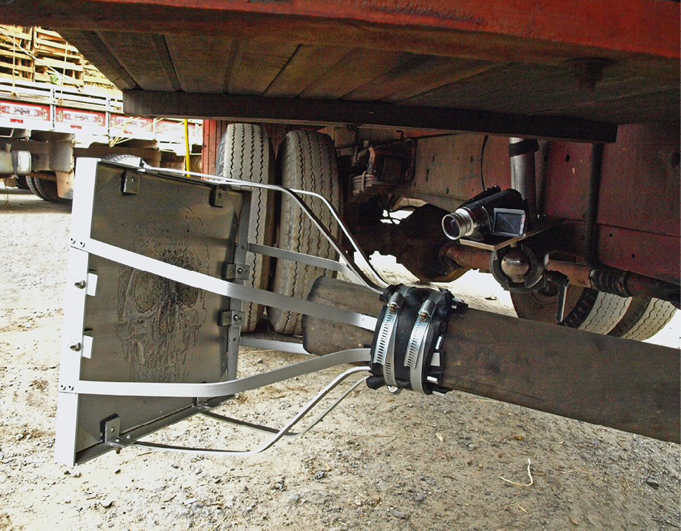 Fotografia. Na parte debaixo de um caminhão, destaque de uma chapa metálica presa por finas vigas de mesmo material a uma coluna de madeira.