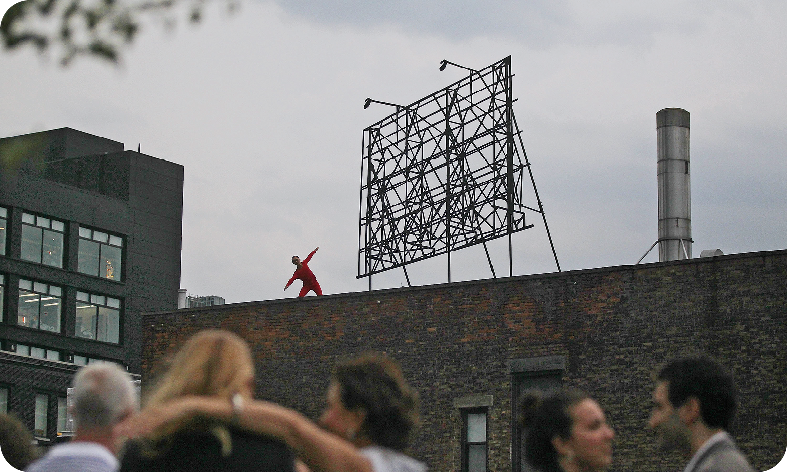 Fotografia. Vista de baixo para cima de uma pessoa que usa roupa vermelha e se movimenta sobre uma plataforma em um telhado em área urbana. Em primeiro plano, há pessoas desfocadas que assistem à performance. Ao fundo, há prédios.