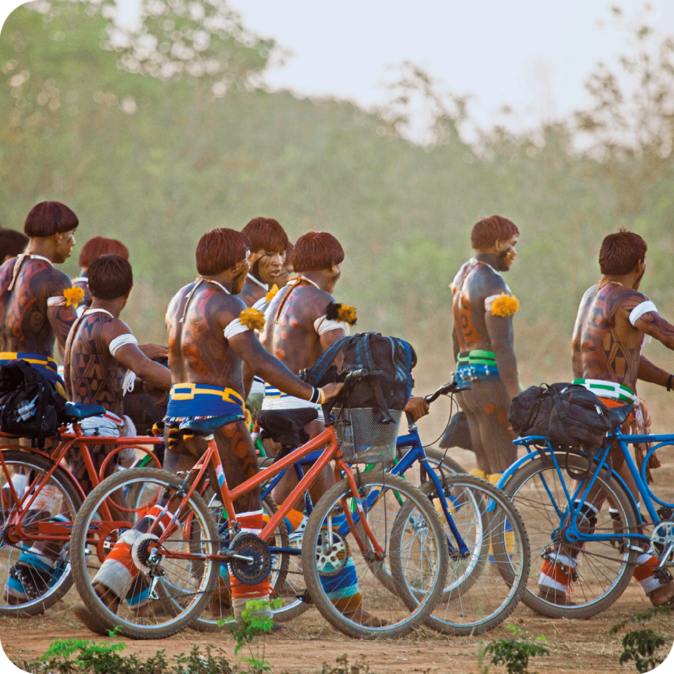 Fotografia. Vista lateral de um grupo de homens indígenas que caminham seminus e empurram bicicletas azuis ou laranjas em uma área de chão de terra. Ao fundo, há algumas árvores.