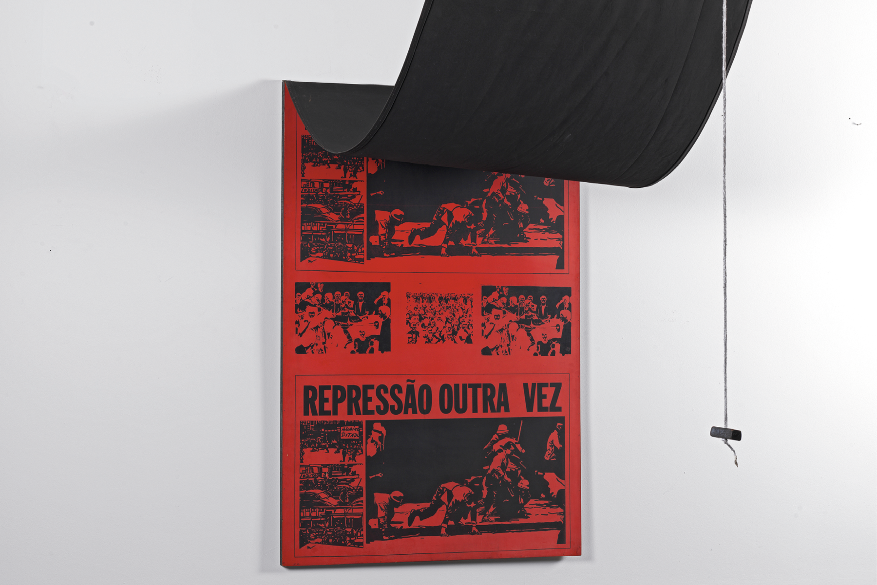 Fotografia. Painel preso à parede com uma cortina levantada sobre ele e que apresenta cor vermelha com quadrinhos em cor preta e onde está escrito REPRESSÃO OUTRA VEZ.