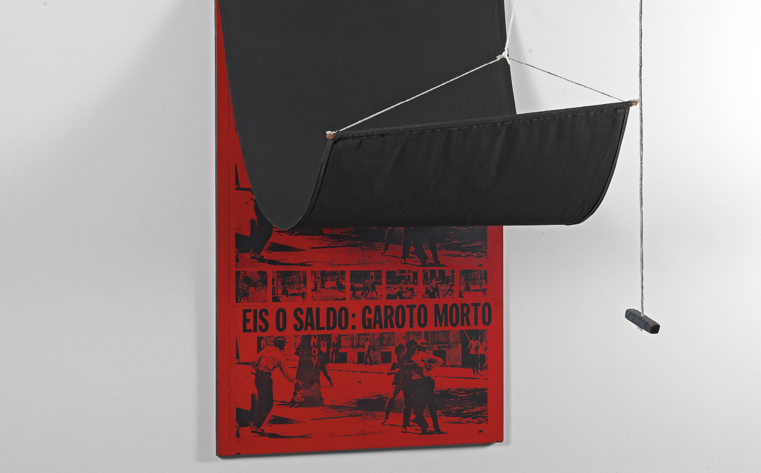 Fotografia. Painel preso à parede com uma cortina levantada sobre ele e que apresenta cor vermelha com quadrinhos em cor preta e onde está escrito EIS O SALDO: GAROTO MORTO.