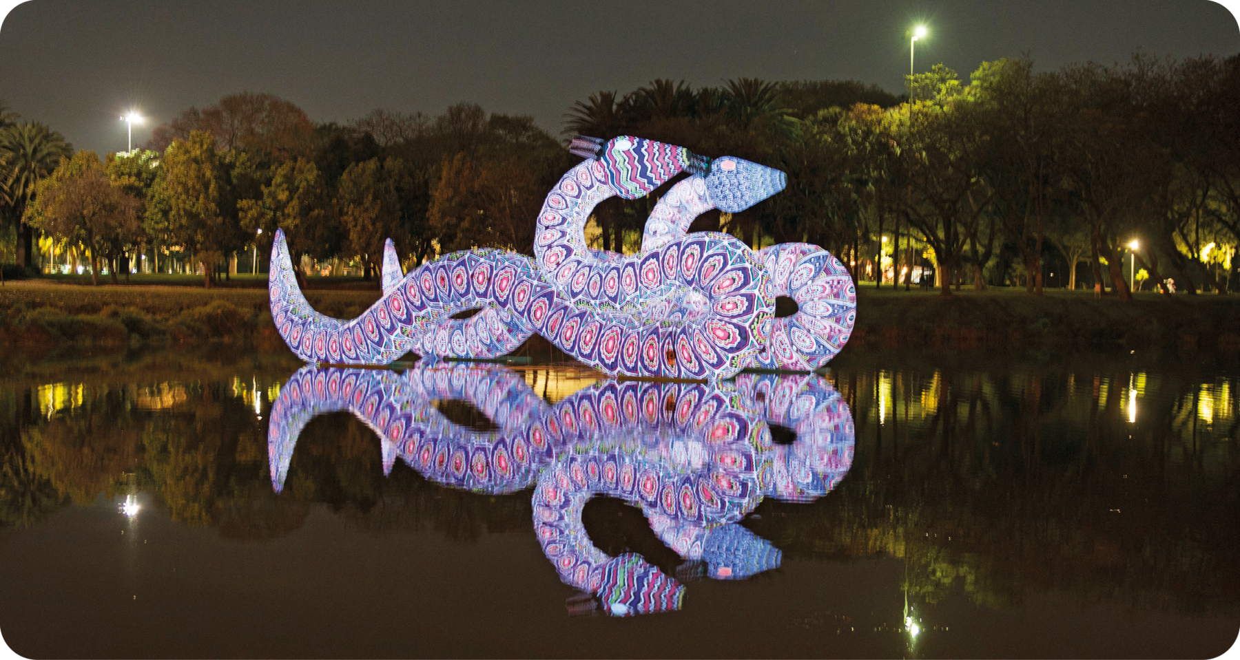 Fotografia. Durante a noite, área que apresenta um lago com estátua de duas serpentes luminosas lado a lado que têm o reflexo projetado na água. Ao fundo, estão árvores e postes de iluminação.
