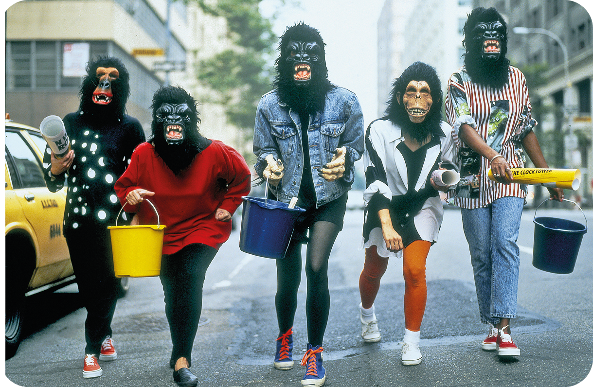 Fotografia. Cinco mulheres caminham lado a lado em uma via usando máscaras de diferentes macacos e seguram baldes e rolos de cartazes. Ao fundo, há prédios.