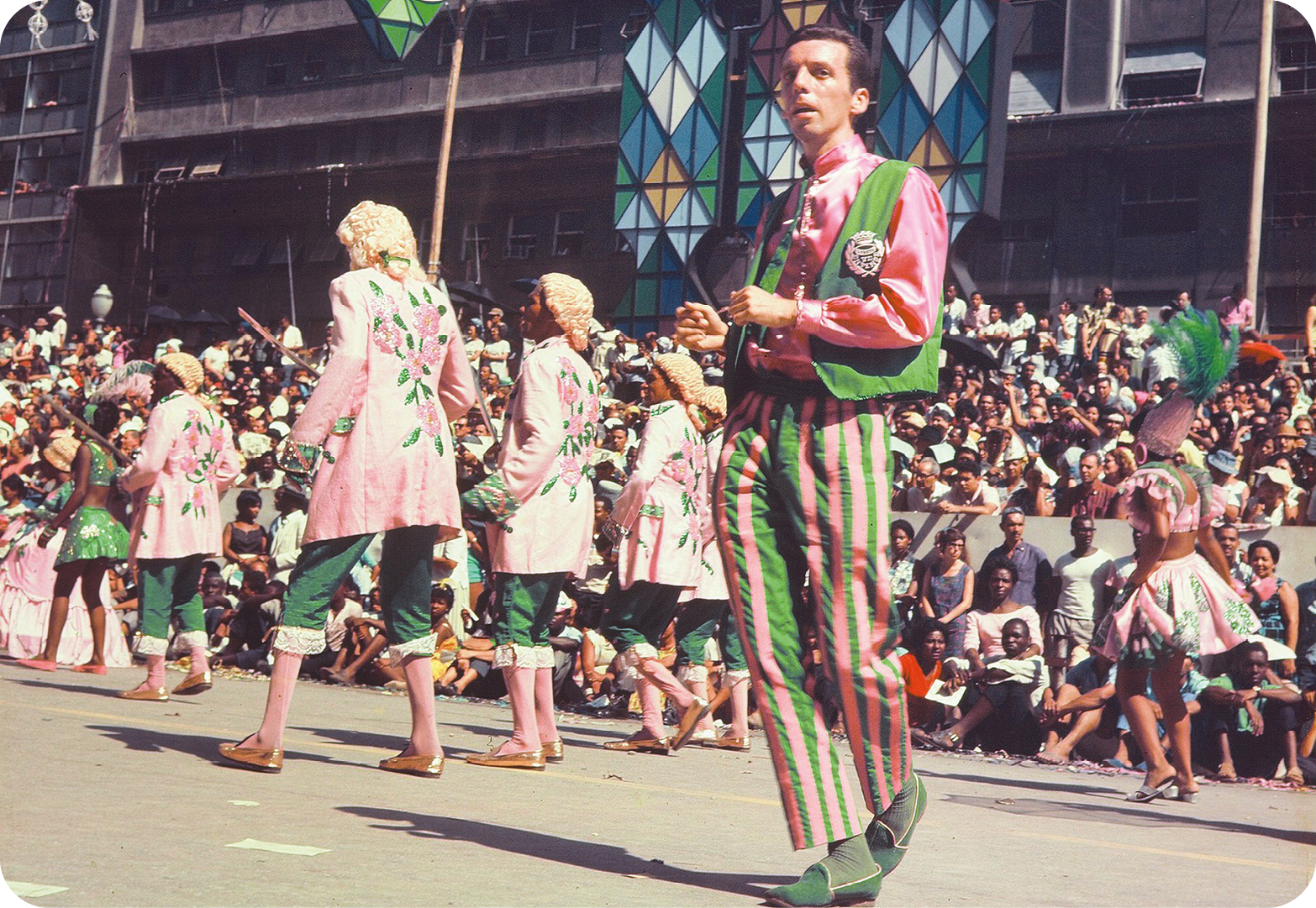 Fotografia. Em primeiro plano, destaque de um homem vestindo figurino verde e rosa que dança em uma via. Em segundo plano, outras pessoas com figurino diferente, mas da mesma cor, desfilam. Ao fundo, uma multidão as assiste.