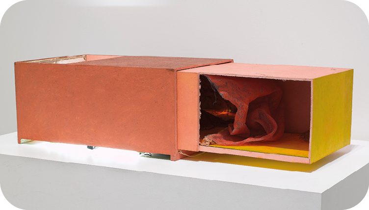 Escultura. Sobre um suporte retangular branco, há uma caixa retangular laranja da qual sai uma outra caixa nas cores amarela e rosa que apresenta uma parte frontal vazada, por onde se vê uma espécie de tecido colorido dentro.