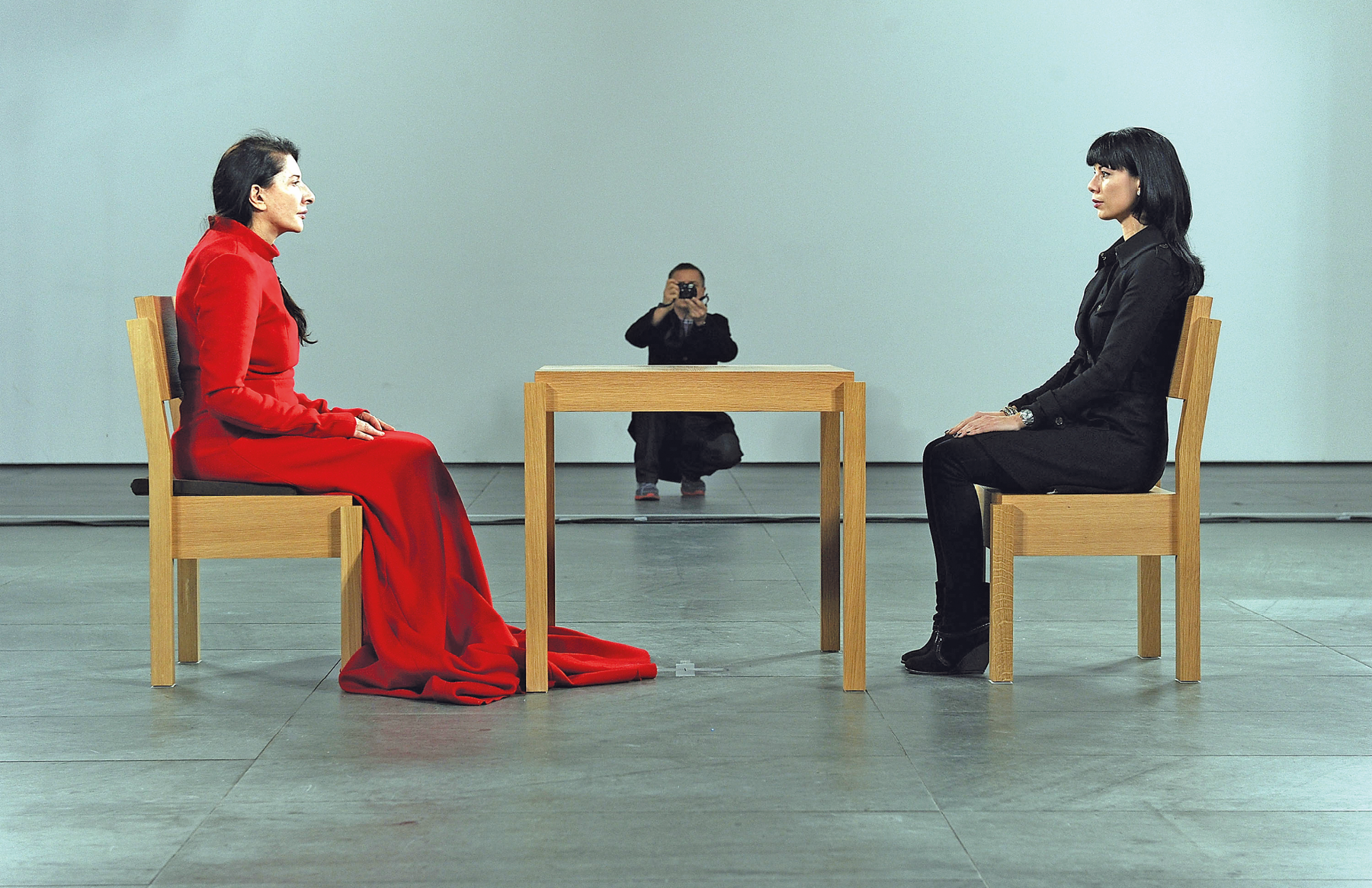 Fotografia. Em uma ampla sala, duas mulheres estão frente a frente sentadas diante de uma mesa. Os móveis são de madeira marrom. A mulher à esquerda veste um longo vestido vermelho; à da direita, usa um traje formal preto. Ao fundo, diante de uma parede acinzentada, há uma pessoa agachada e fotografando as mulheres.