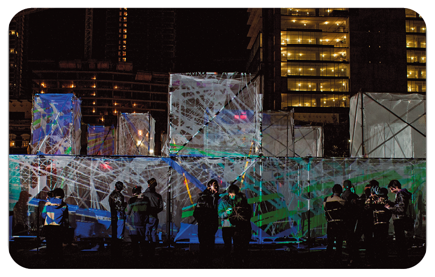 Fotografia. Durante a noite, em um ambiente aberto, um grupo de pessoas está ao redor de um conjunto de cubos com superfície transparente com projeção de linhas coloridas. Ao fundo, prédios com luzes acesas.