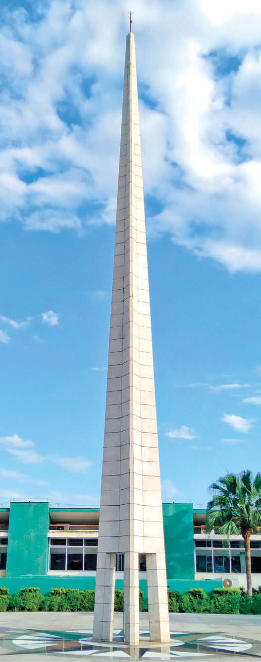 Fotografia. Destaque de uma área aberta onde há um obelisco revestido por mármore branco. Ao fundo, há um prédio e o céu azul.