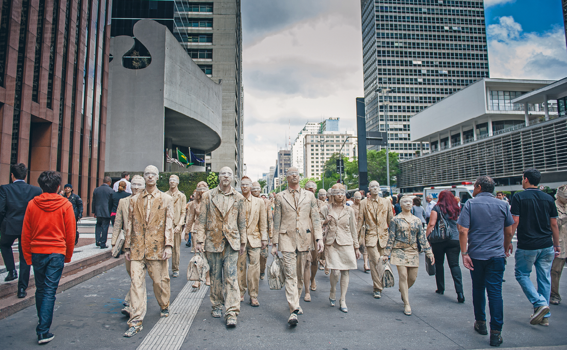 Fotografia. Em uma via urbana rodeada por prédios, um grupo de pessoas com corpo e roupas formais pintados de bege caminha na mesma direção e são vistos de frente. Ao redor, transeuntes passam em sentido contrário.