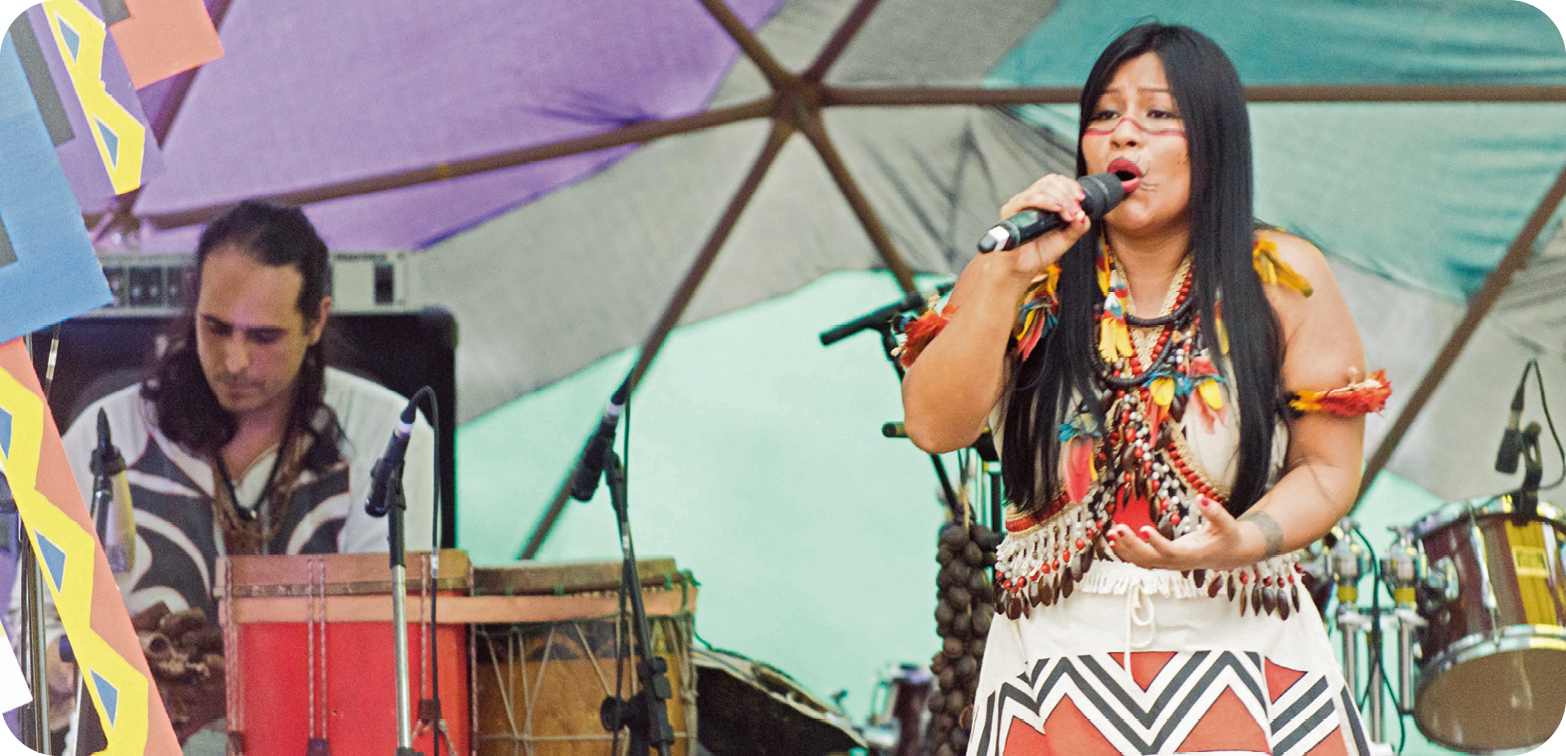 Fotografia. À direita, destaque de uma mulher indígena que canta ao microfone. Ela tem cabelo longo, liso e preto, penteado para o lao, o rosto e os braços pintados em linhas horizontais e adereços no pescoço, tronco e braços. Ao fundo, à esquerda, um rapaz toca percussão.