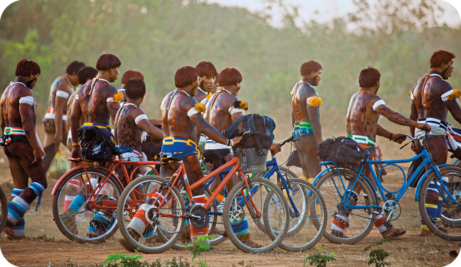 Fotografia. Vista lateral de um grupo de homens indígenas que caminham seminus e empurram bicicletas azuis ou laranjas em uma área de chão de terra. Ao fundo, há algumas árvores.