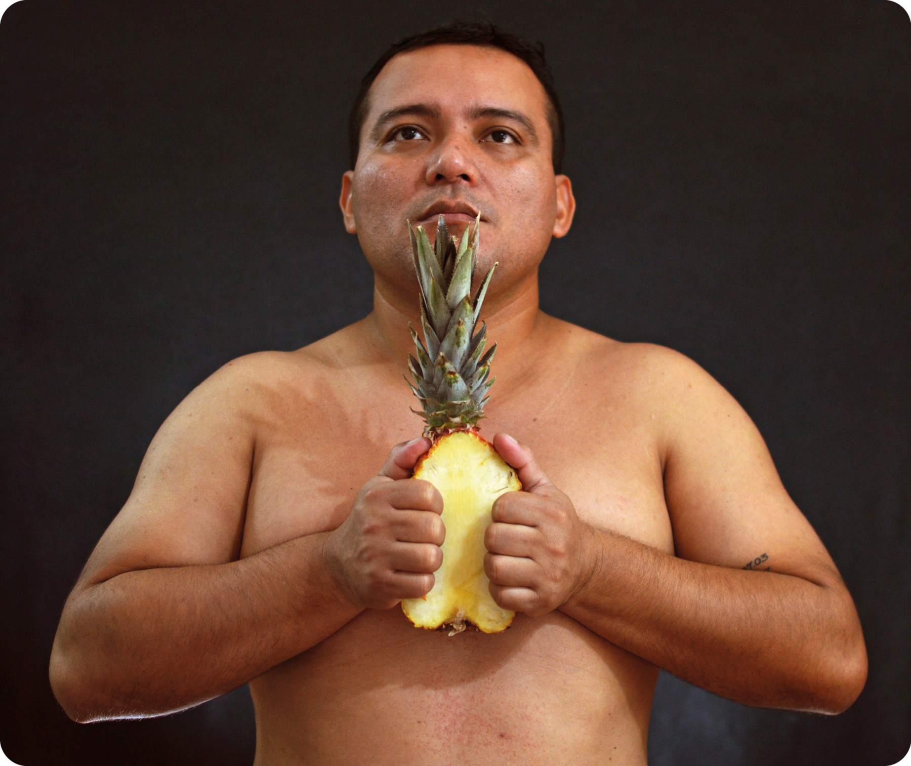 Fotografia. Retrato de um homem sem camisa visto da cintura para cima à frente de um fundo preto. Ele tem sobrancelhas e lábios grossos, nariz largo e segura um abacaxi cortado ao meio, o qual está com a polpa virada para frente.
