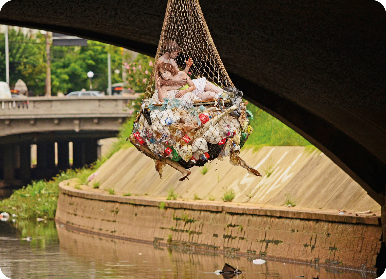 Fotografia. Plano aberto que apresenta uma rede de pesca repleta de lixo pendurada em uma ponte acima de um rio com dejetos. Acima do lixo e dentro da rede, há um casal. Ao fundo, trecho urbano.