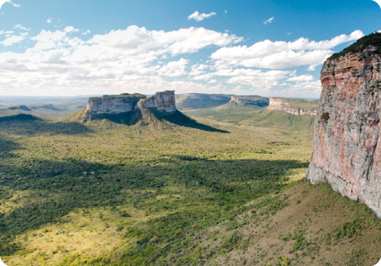 Fotografia. Vista de uma área extensa com vegetação e grandes montanhas rochosas. O céu é azul-claro com nuvens brancas.