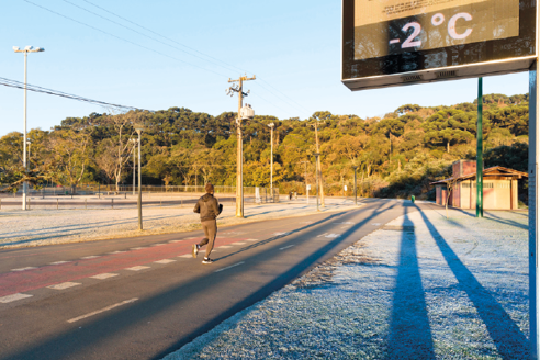 Fotografia. Uma pessoa correndo em meio a uma rua. Nas laterais, grama encoberta por uma camada branca de gelo; ao fundo, árvores e postes com fios. No canto superior direito, um visor marcando menos 2 graus.