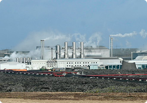 Fotografia com uma grande construção com chaminés que expelem fumaça e grandes estruturas tubulares de metal. Ao redor, vegetação.