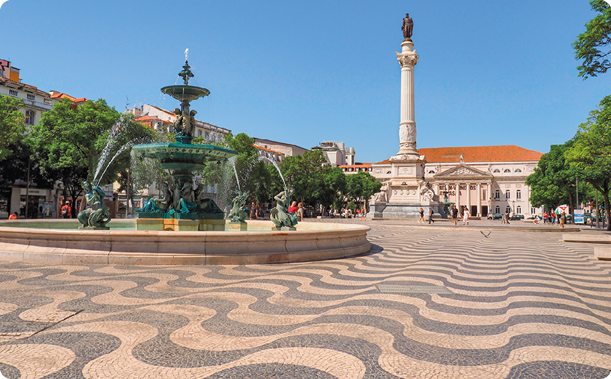 Fotografia. Praça com chão composto por piso feito com pequenos pedaços de pedras que formam linhas curvas brancas e pretas. Ao centro, uma fonte com estátuas; ao fundo, grande construção com torre e estátua no topo. Há pessoas e árvores ao redor.