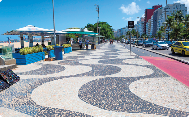 Fotografia. Calçada com chão composto por piso feito com pequenos pedaços de pedras que formam linhas curvas brancas e pretas. À esquerda, barracas; à direita, rua com carros e prédios.