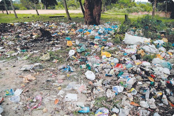 Fotografia. Vista de um local com muitos resíduos espalhados pelo solo, dentre eles, plástico, papel e outros materiais. No entorno, há grama e árvores.