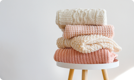 Fotografia. Quatro blusas de lã dobradas e colocadas sobre um banco. Duas são brancas e as outras são rosa.