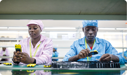 Fotografia. À esquerda, mulher usando touca e uniforme cor-de-rosa, segurando um aparelho eletrônico em frente a uma esteira. À direita, homem usando touca e uniforme azul, também em frente à esteira, segurando um aparelho eletrônico sobre produtos. 