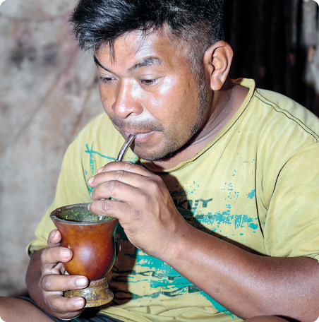 Fotografia. Um homem, representado da cintura para cima, segurando uma cuia com conteúdo verde e com um canudo de metal na boca.