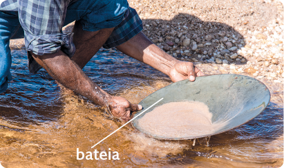 Fotografia. Um homem agachado em meio a um corpo d'água, segurando uma bateia, recipiente plano e redondo, que está com terra.