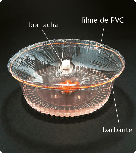 Fotografia. Uma bacia plástica com líquido dentro. Há um filme de PVC cobrindo a parte superior da bacia, preso por um barbante. No centro, uma borracha quadrada e branca.