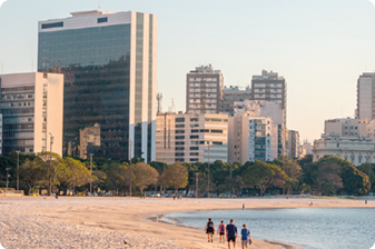 Fotografia. Vista de pessoas andando pela areia de uma praia. Ao fundo, árvores e prédios.