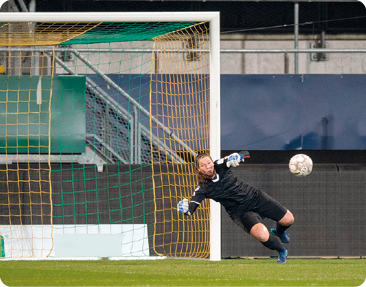 Fotografia. Uma mulhervestindo um uniforme preto e luvas grossas está curvada para o lado, com os braços estendidos para frente. Ela está sobre um gramado com as traves de um gol de futebol atrás. Uma bola está no ar em direção a ela.