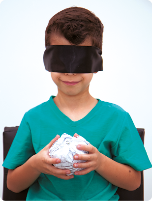 Fotografia. Uma criança sentada, representada da cintura para cima, segurando com as duas mãos uma esfera feita de papel. Ele está sorrindo e com os olhos vendados.