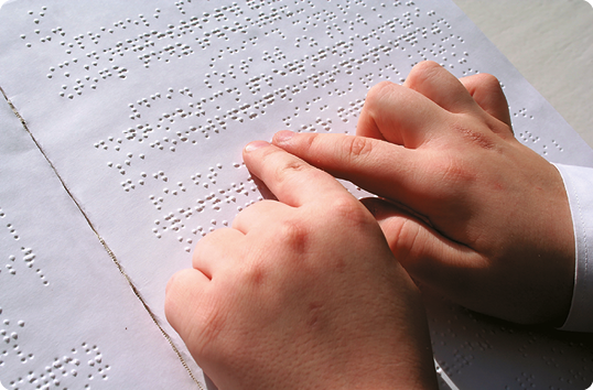 Fotografia. As mãos de uma criança estão sobre uma folha em branco, onde há escrita em Braille com pontos em relevo.