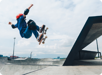 Fotografia. Uma criança em um skate saltando. Ela usa capacete, joelheira e cotoveleira. Está agachada, com os dois pés sobre o skate no ar e os braços levemente esticados e levantados. À direita e abaixo, uma rampa. Ao fundo, outras rampas.