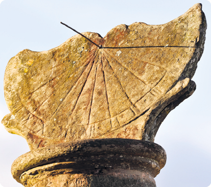 Fotografia. Escultura de pedra de um relógio de sol. Estrutura achatada sobre uma base e com uma haste vertical ao centro que faz sombra à direita sobre a estrutura achatada.